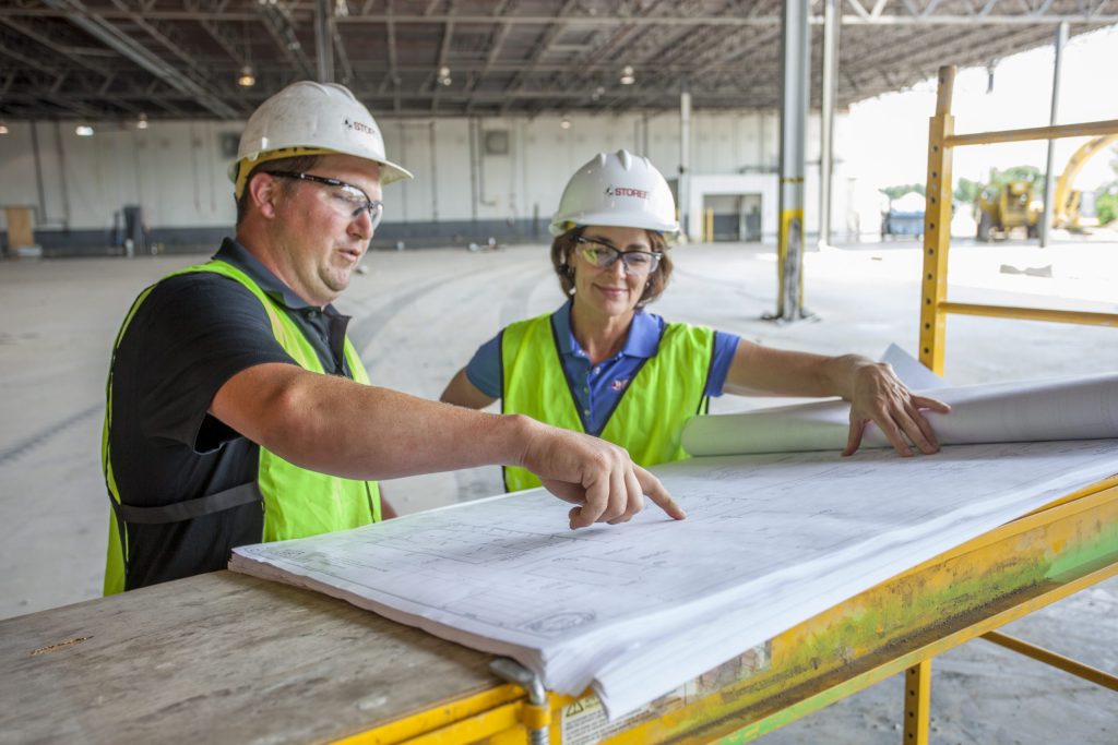 A man and woman in hard hats and safety vests look over plans at a construction site.