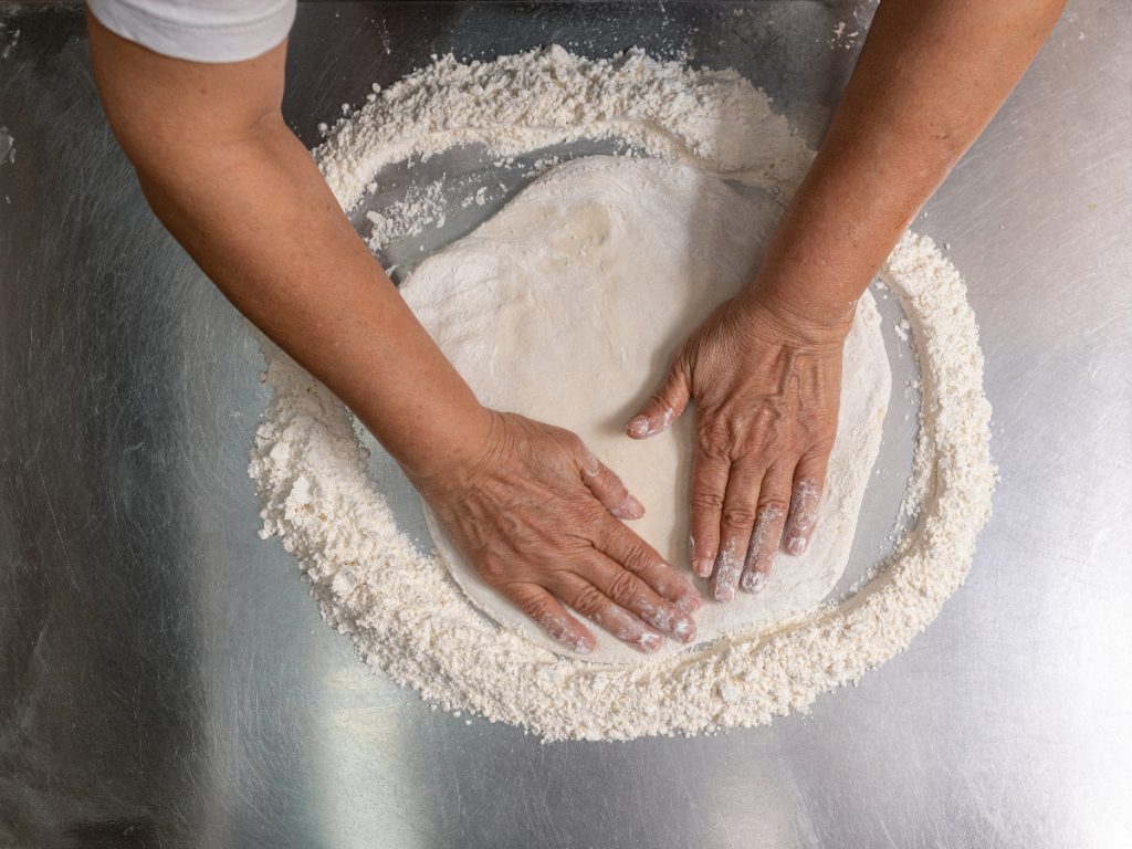 Two hands kneading pizza dough on a stainless steel table to illustrate work table fabrication.